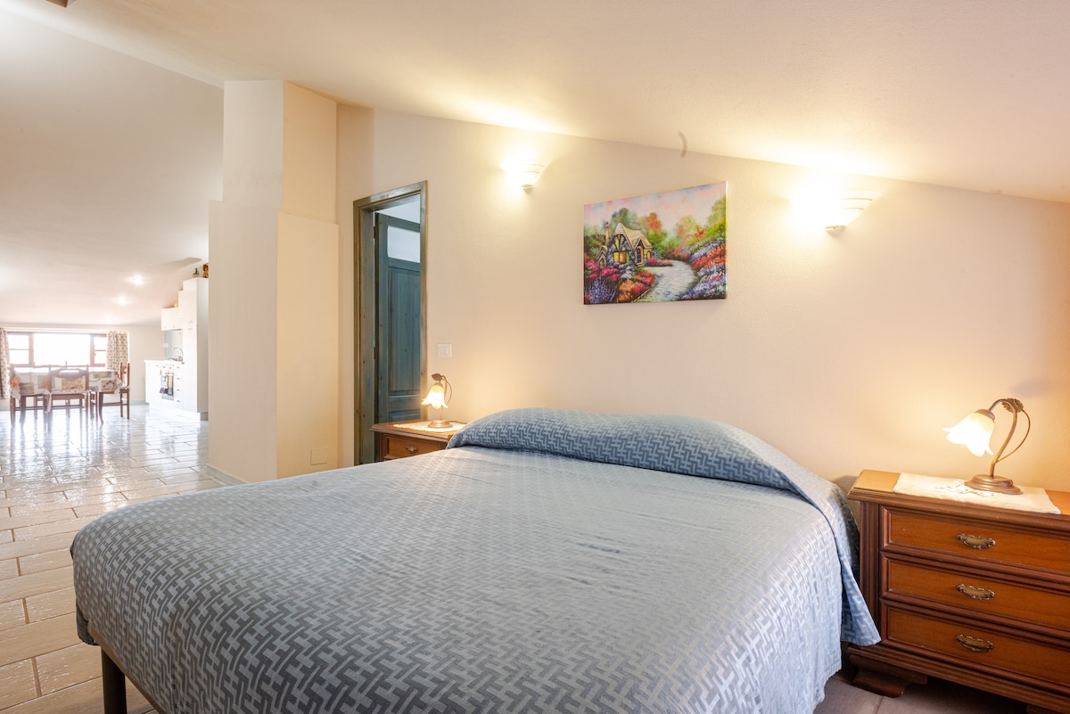 A spacious sleeping area features a large bed with a patterned blue coverlet. Each bedside table is equipped with a lamp, providing soft lighting. A colorful artwork hangs above the bed, and natural light filters in from a nearby window.
