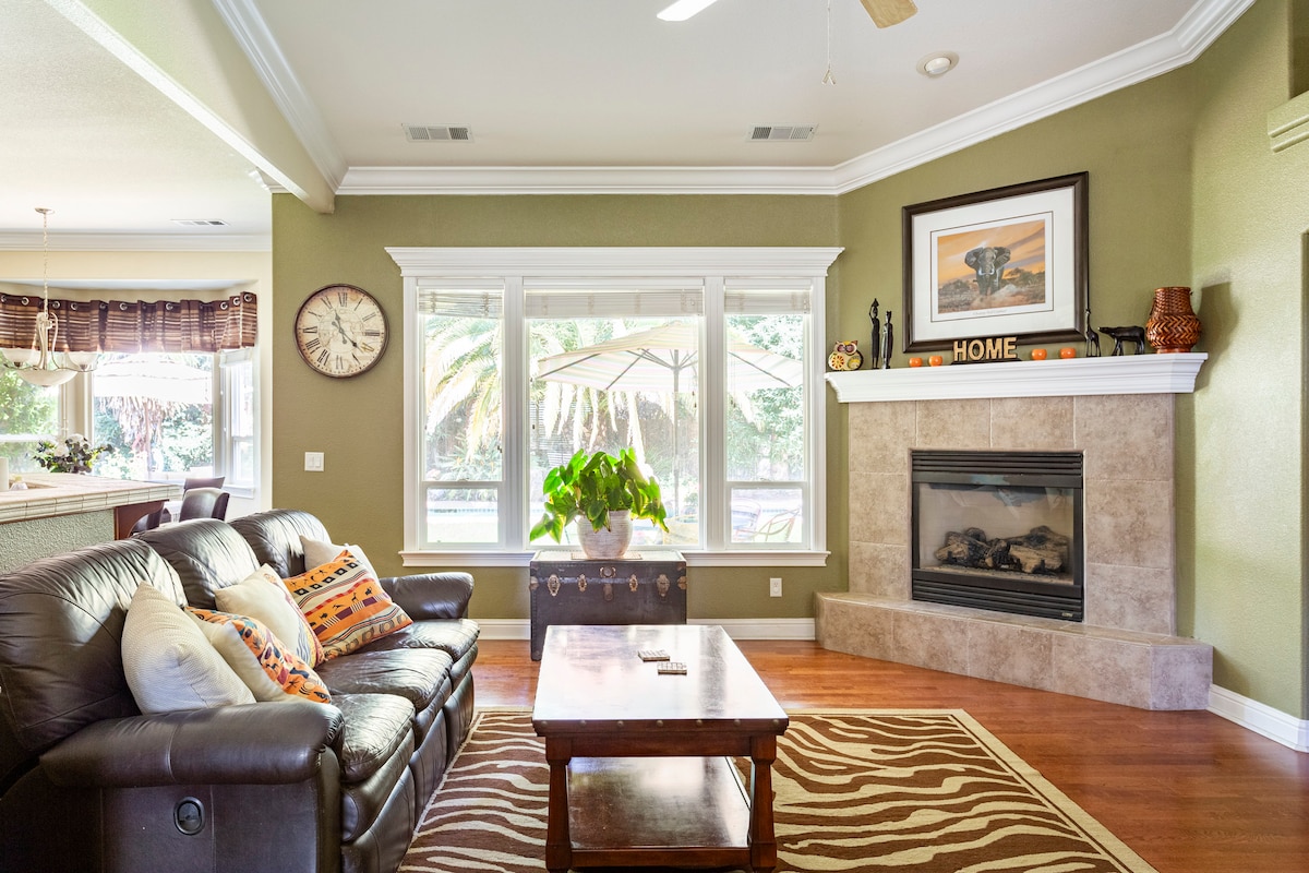 A cozy living room features a dark leather sofa adorned with patterned cushions, positioned near a stylish coffee table. A warm fireplace is framed by light stone, and large windows provide a view of lush greenery outside. Elegant accents and a large clock enhance the space.