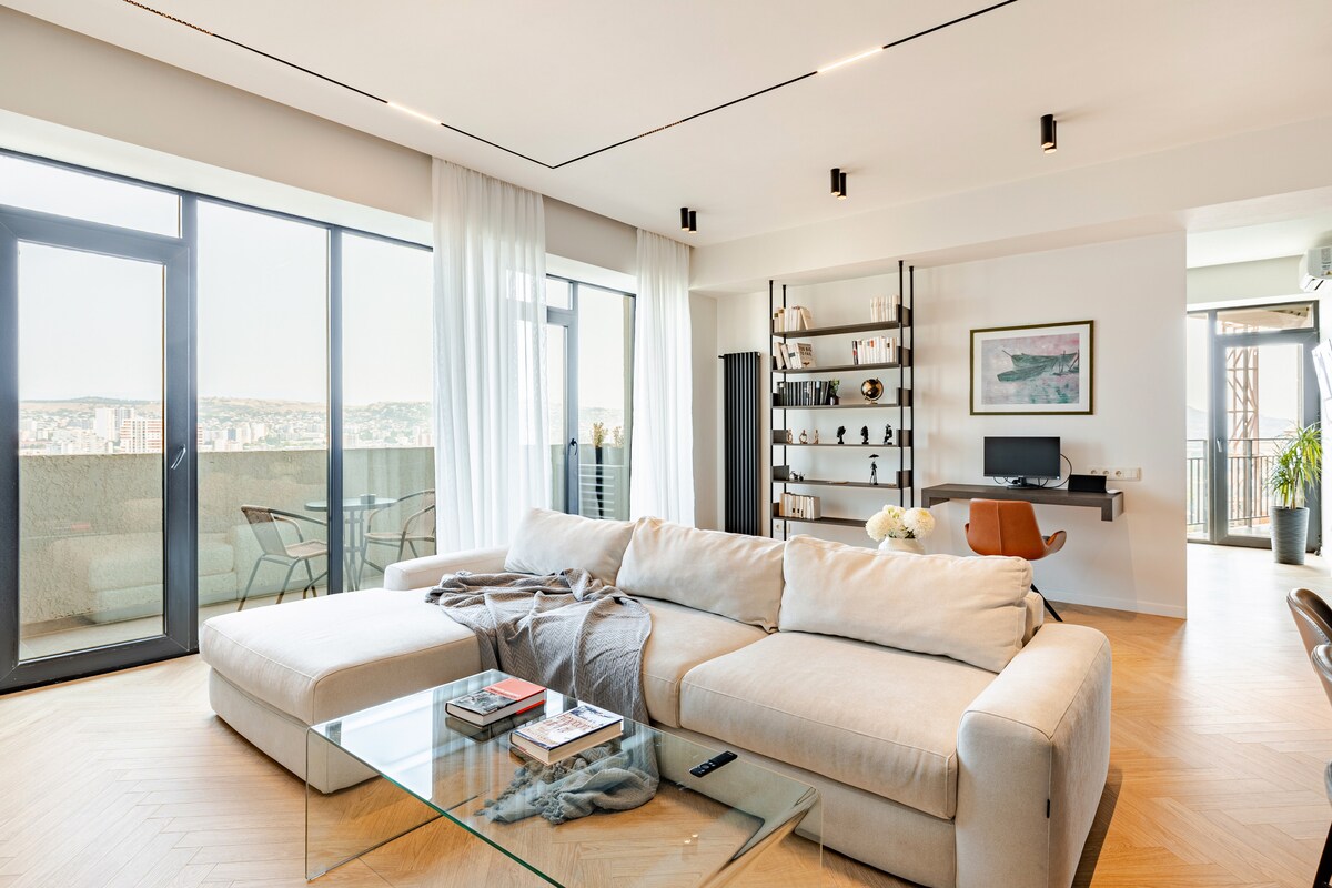 A spacious living room is depicted with a light-colored sectional sofa facing a glass coffee table. Large windows provide ample natural light and showcase views of the city. A contemporary desk setup is visible, accompanied by a bookshelf in the background.