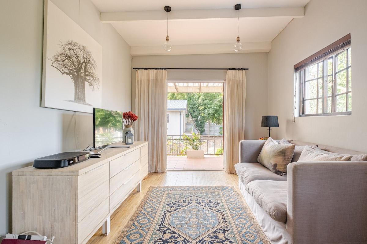 An open-plan living area features a soft couch and a wooden TV console with a flat-screen television. Natural light pours in through large sliding doors leading to an outdoor area, while a decorative area rug adds warmth to the space.