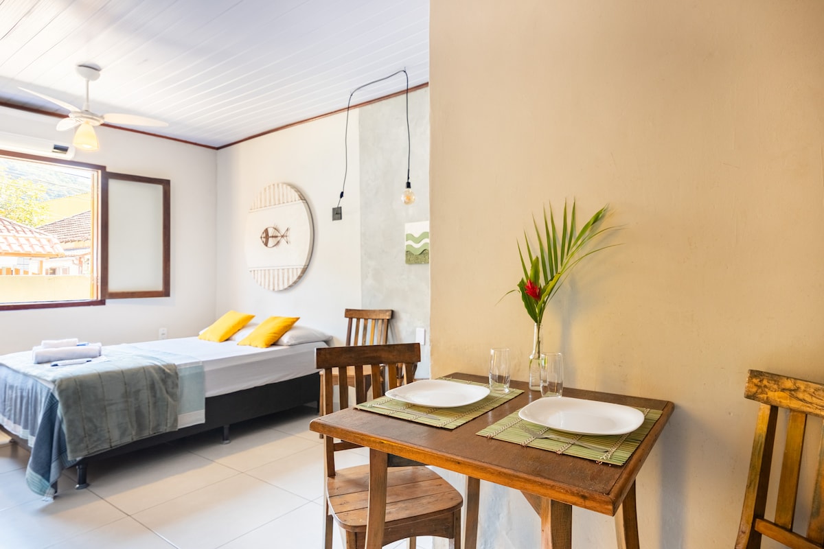 A cozy area includes a wooden table set for two, featuring plates and glasses. A vibrant flower is placed in the center. The sleeping area features a spacious bed with yellow pillows, and natural light enters through a window with a wooden frame.