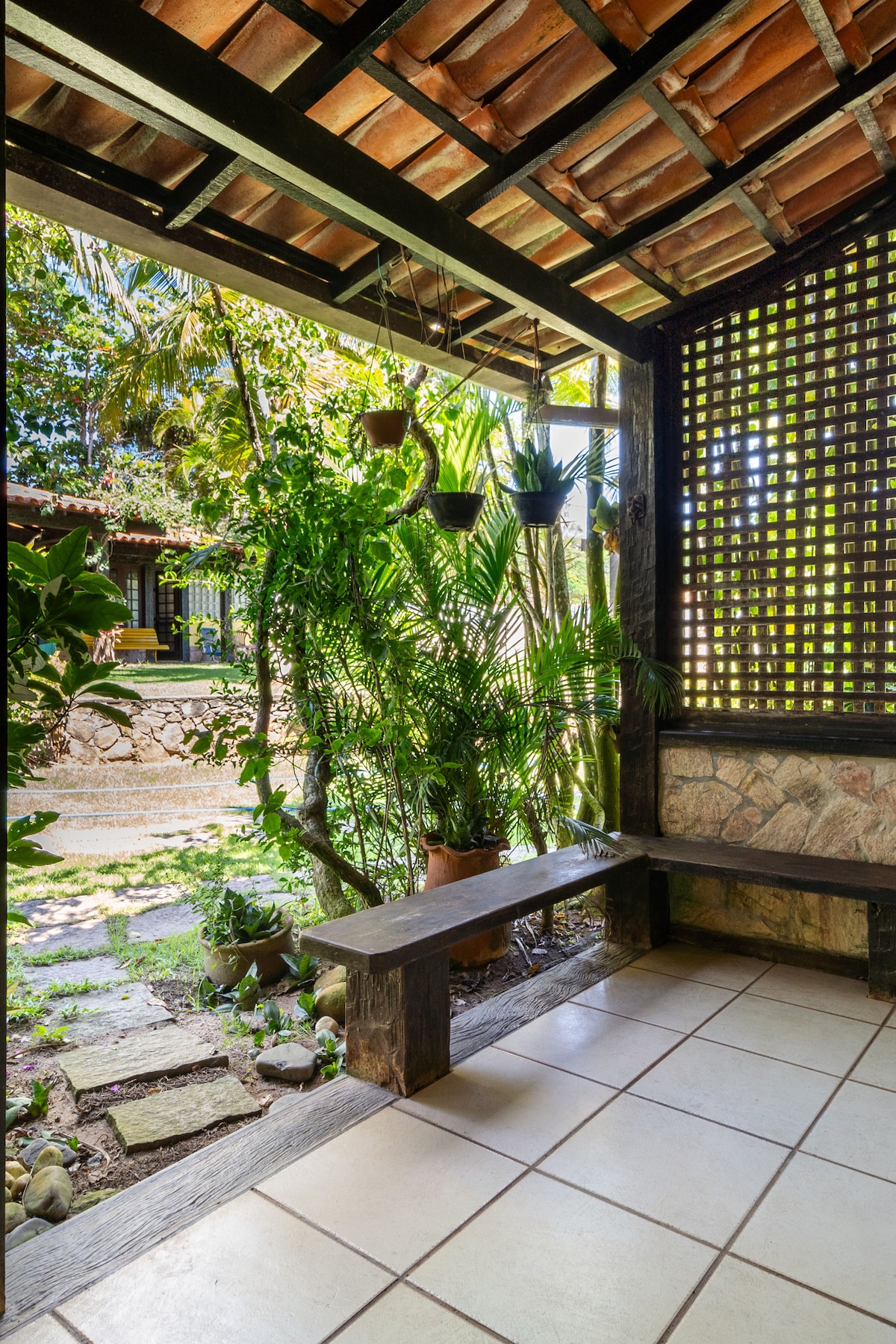 A shaded entryway features stone flooring and a wooden bench. Lush greenery, including hanging plants and potted vegetation, surrounds the space, creating a serene atmosphere. Natural light filters through the lattice work, enhancing the connection to the outdoors.
