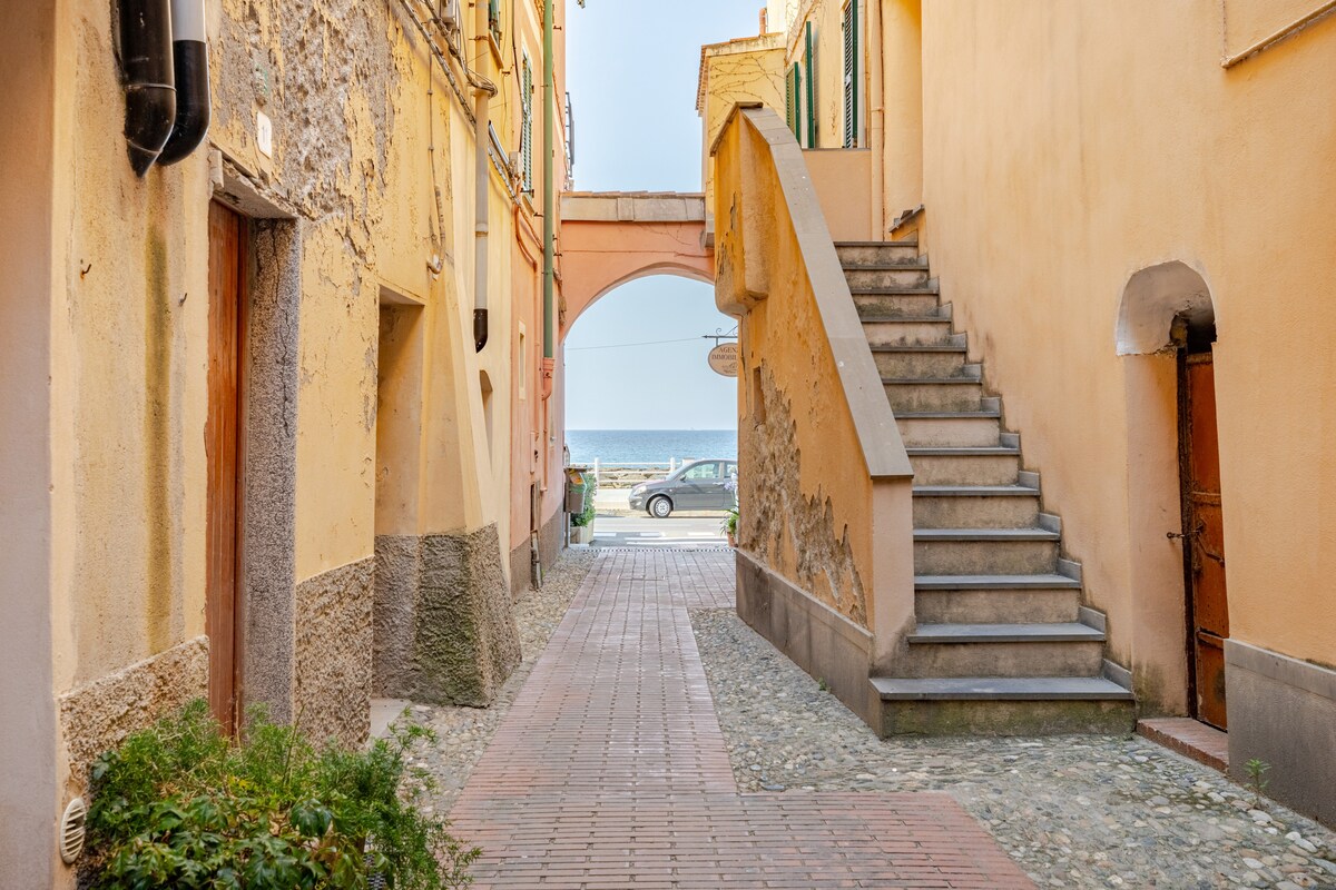 A serene street scene is depicted, featuring a cobblestone path that leads to an archway. On the left, a staircase ascends alongside colorful building facades. The sea can be seen in the distance, providing a glimpse of the coastal location.