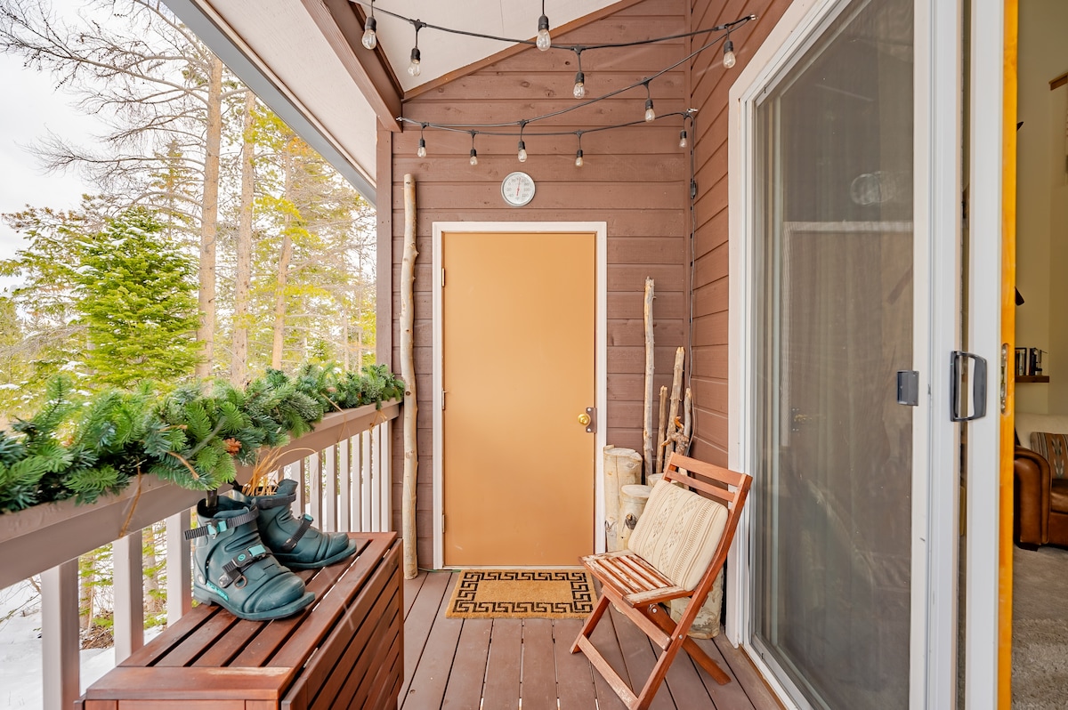 A welcoming entrance features a wooden deck adorned with string lights. A decorative garland complements the space, while a pair of boots rests beside a folding chair. The central door, marked with a clock, is framed by glass doors leading indoors.