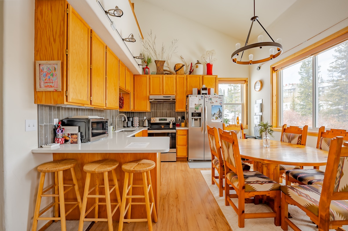A functional kitchen area features wooden cabinetry and modern appliances. A large dining table is surrounded by comfortably cushioned chairs, with natural light streaming in from windows that overlook the serene outdoor space.