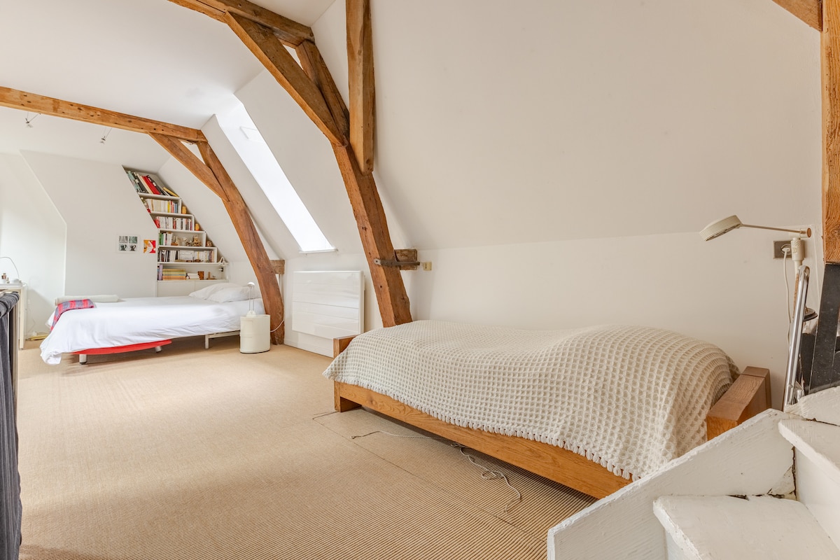 A spacious loft area is characterized by exposed wooden beams. A double bed is positioned alongside a single bed, both topped with cozy bedding. Natural light filters through a nearby window, illuminating the light-colored walls and wooden accents, creating an open feel.