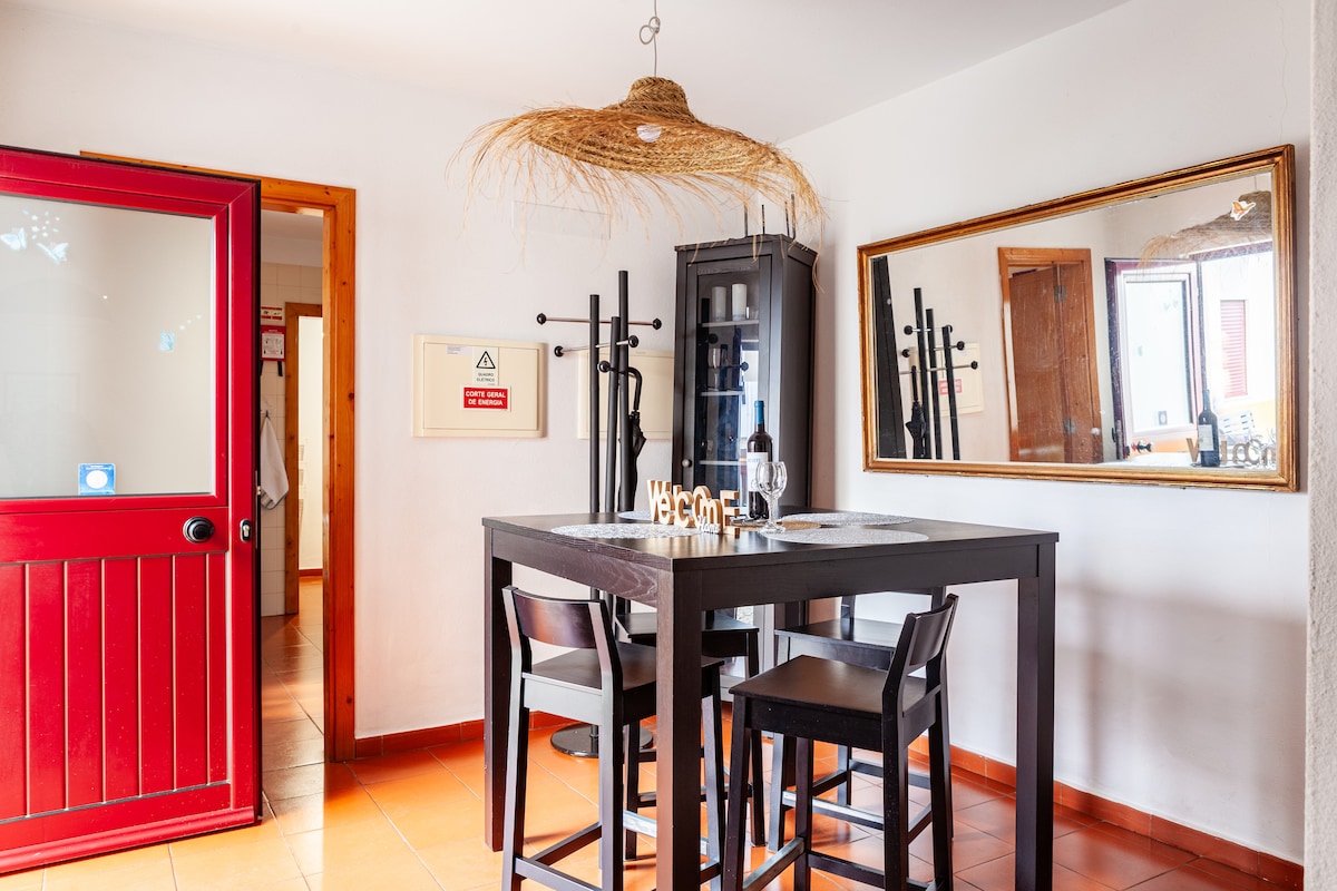 A dining area features a tall, dark wooden table accompanied by four high-backed chairs. A stylish pendant light hangs above, and a mirror reflects the space. A decorative welcome sign and natural elements add warmth to the inviting setting.