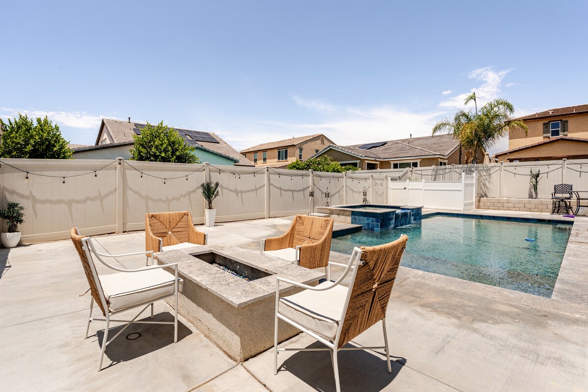 An outdoor area features a pool surrounded by a concrete deck and decorative fencing. Four chairs are positioned around a fire pit, creating a casual space for relaxation. Lush greenery and a palm tree add a touch of nature, with sunny skies overhead.