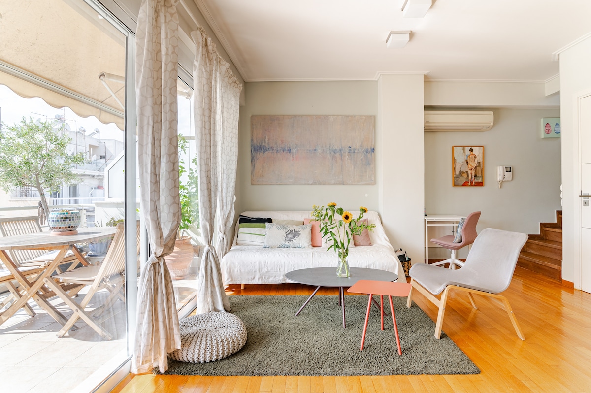 A bright and inviting living room features a cozy sofa bed adorned with colorful cushions. A stylish coffee table is centered on a soft rug, surrounded by modern seating. Large glass doors provide access to the outdoor terrace, enhancing the airy atmosphere.