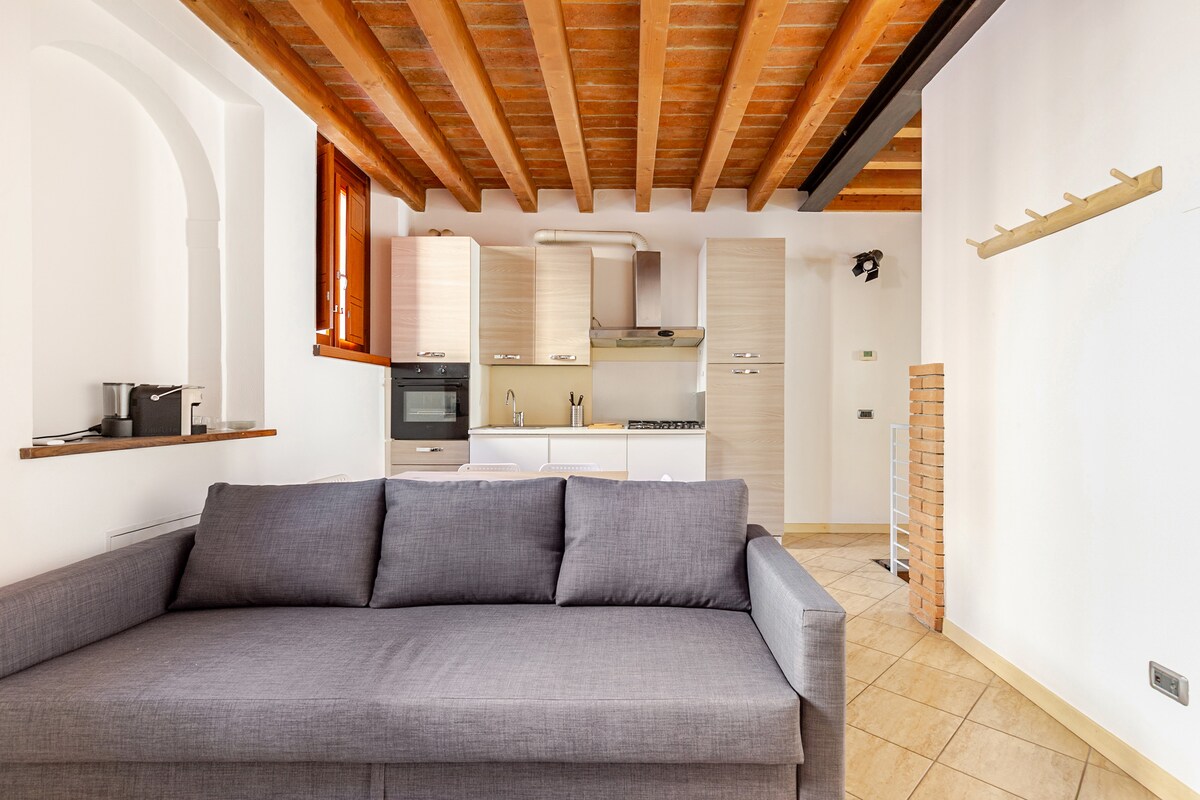 A modern kitchen area features light wood cabinets and stainless steel appliances. A gray sofa provides comfortable seating, situated near a window that allows natural light to illuminate the space. Exposed wooden beams enhance the visual interest of the ceiling.