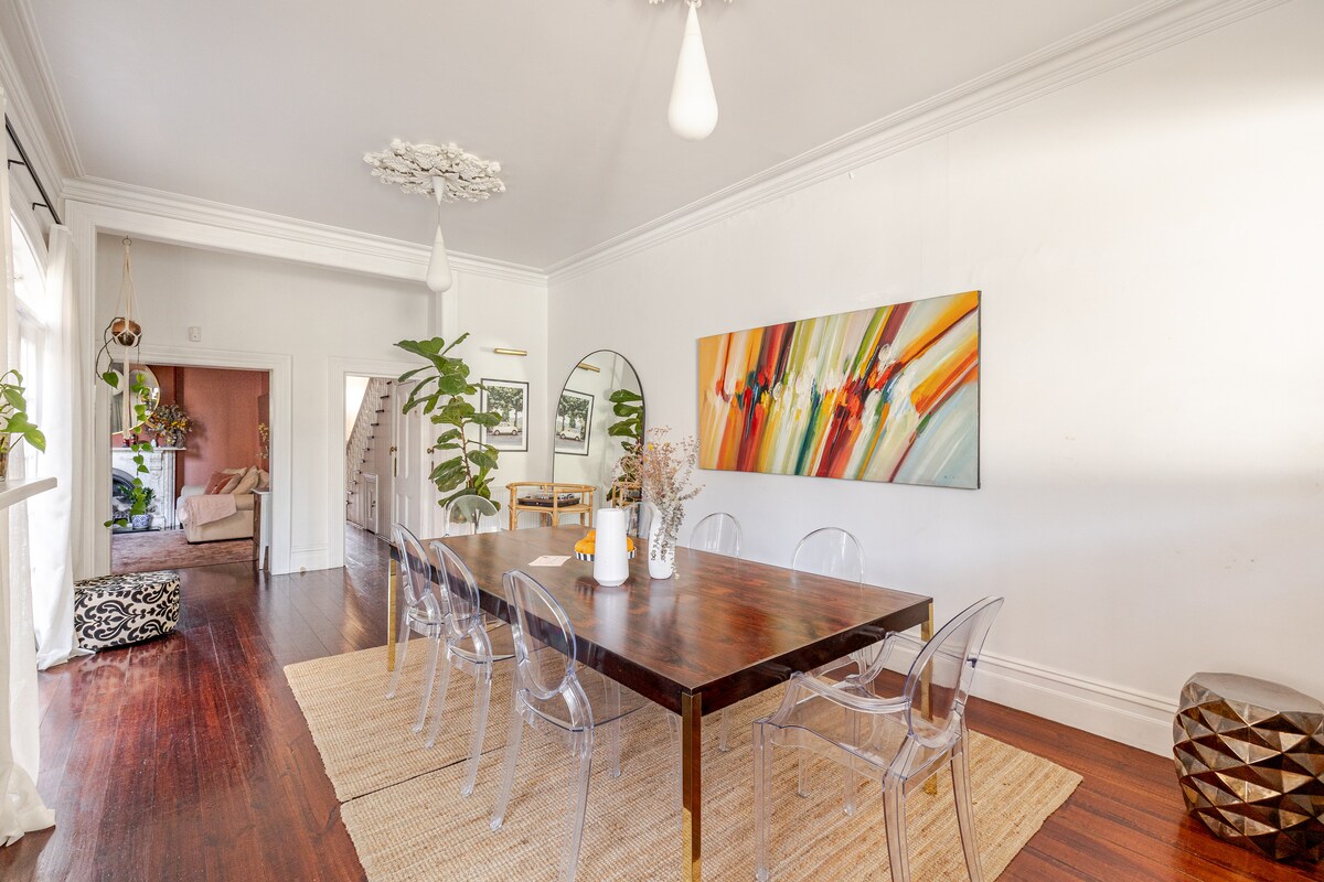 A spacious dining area is highlighted by a large wooden table surrounded by transparent chairs. Light from multiple windows brightens the room, complemented by indoor plants and a vibrant artwork on the wall. A textured rug adds warmth to the polished wooden floor.