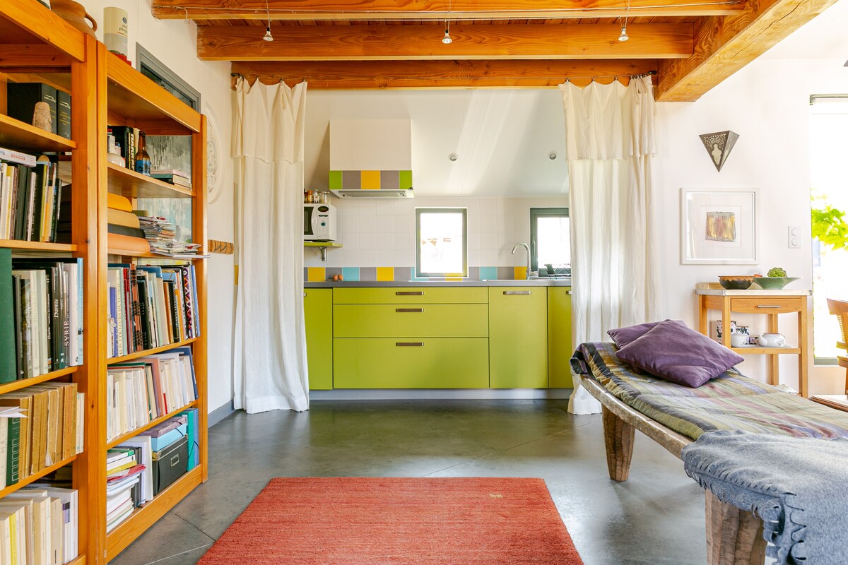 The open living space is characterized by bright green cabinetry and a modern kitchen setup. Wooden beams add warmth to the ceiling. A wooden bookshelf filled with books stands against one wall, while a cozy seating area features a simple bench and a colorful area rug.