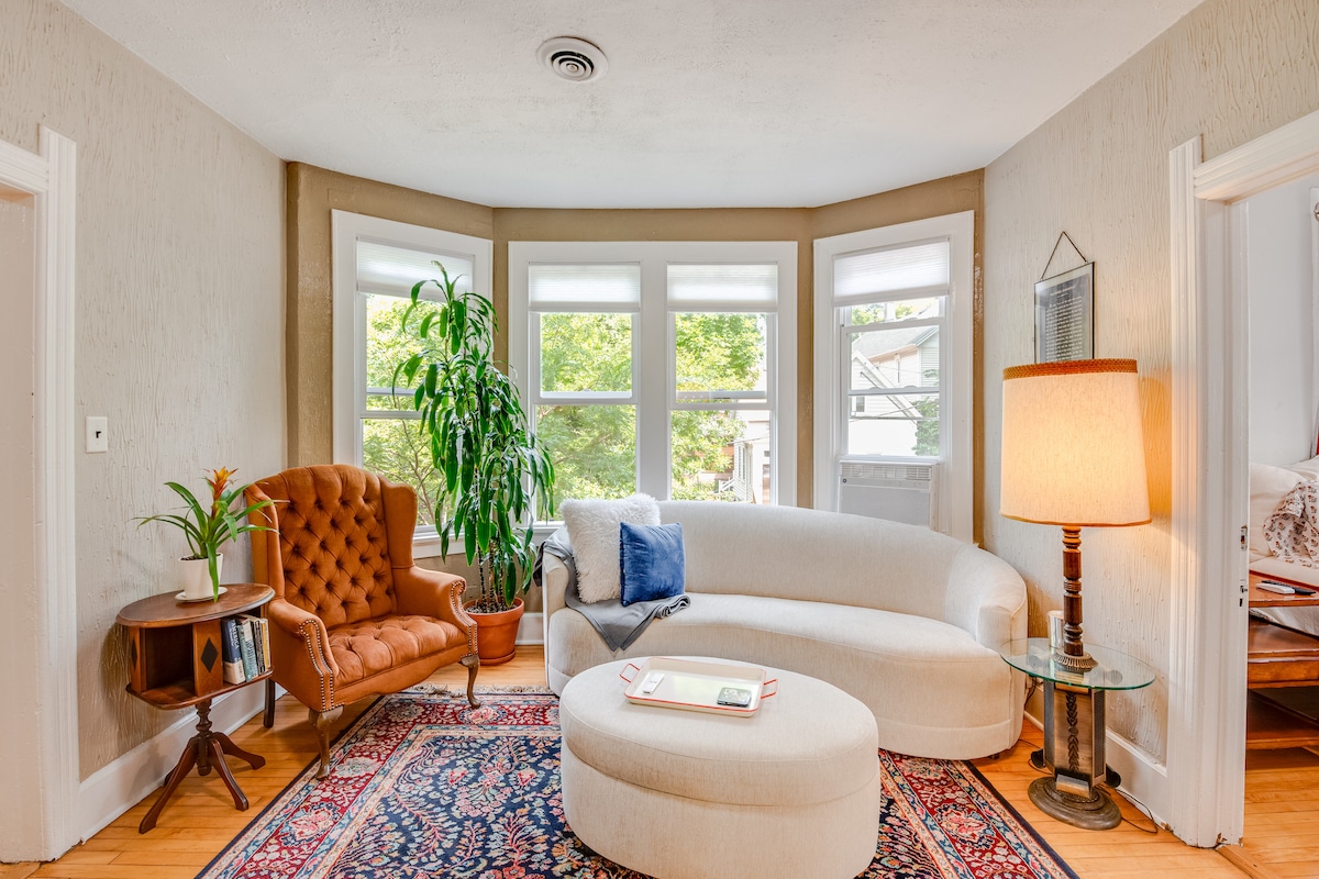A cozy living area is filled with natural light, featuring a curved white sofa and a brown upholstered chair. A round coffee table sits in the center, while a large potted plant and decorative elements add a touch of warmth. Large windows frame views of the outdoors.