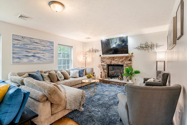 Acogedor y acogedor salón con un gran sofá seccional, asientos de felpa, chimenea de piedra y TV de pantalla plana. Las ventanas luminosas llenan el espacio de luz natural, perfecto para relajarse, ver películas o disfrutar del tiempo con familiares y amigos.