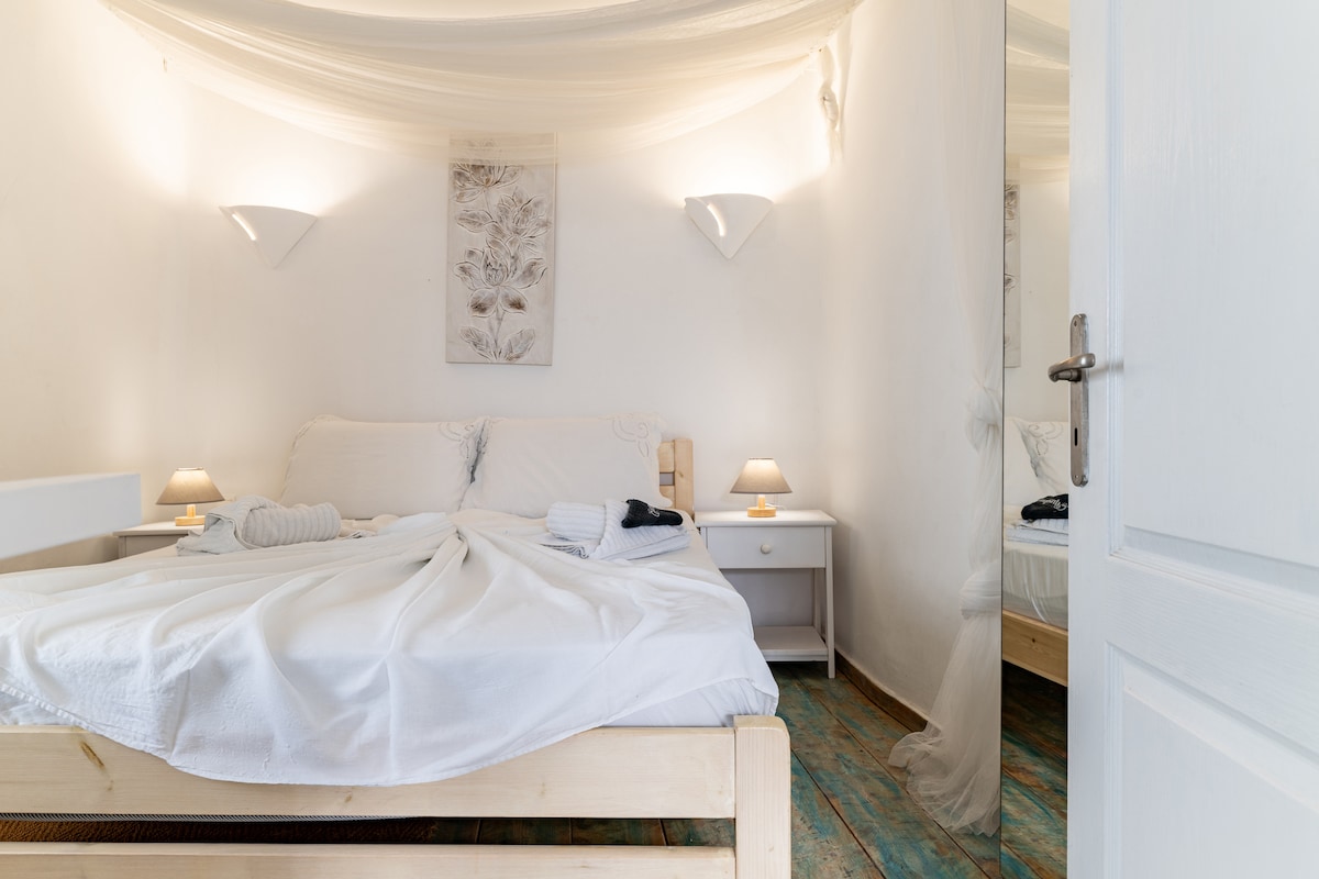 Habitación 1 con aire acondicionado y cama doble romántica (1,40 × 2,00) con armario y ventana hacia una calle tradicional micónica estrecha. Techo alto, ¡esta es la habitación para personas más altas! 