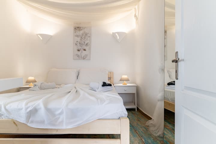Værelse 1 med aircondition og dobbelt romantisk seng (1,40 × 2,00) med skab og vindue mod en traditionel smal mykonisk gade. Højt til loftet er det plads til højere personer !