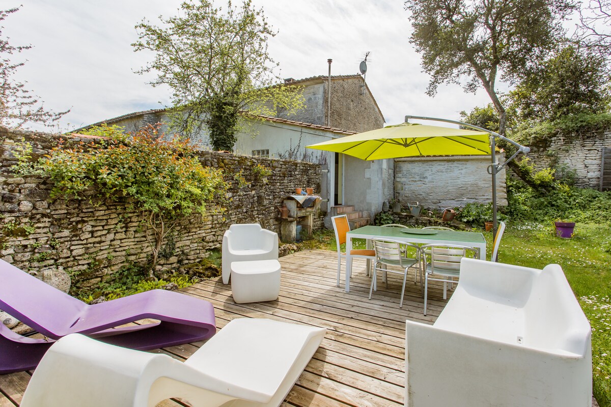 An outdoor terrace features a wooden deck, with a dining table and chairs beneath a bright yellow umbrella. Two lounge chairs in purple and white are positioned nearby, surrounded by greenery and a stone wall that adds privacy to the space.