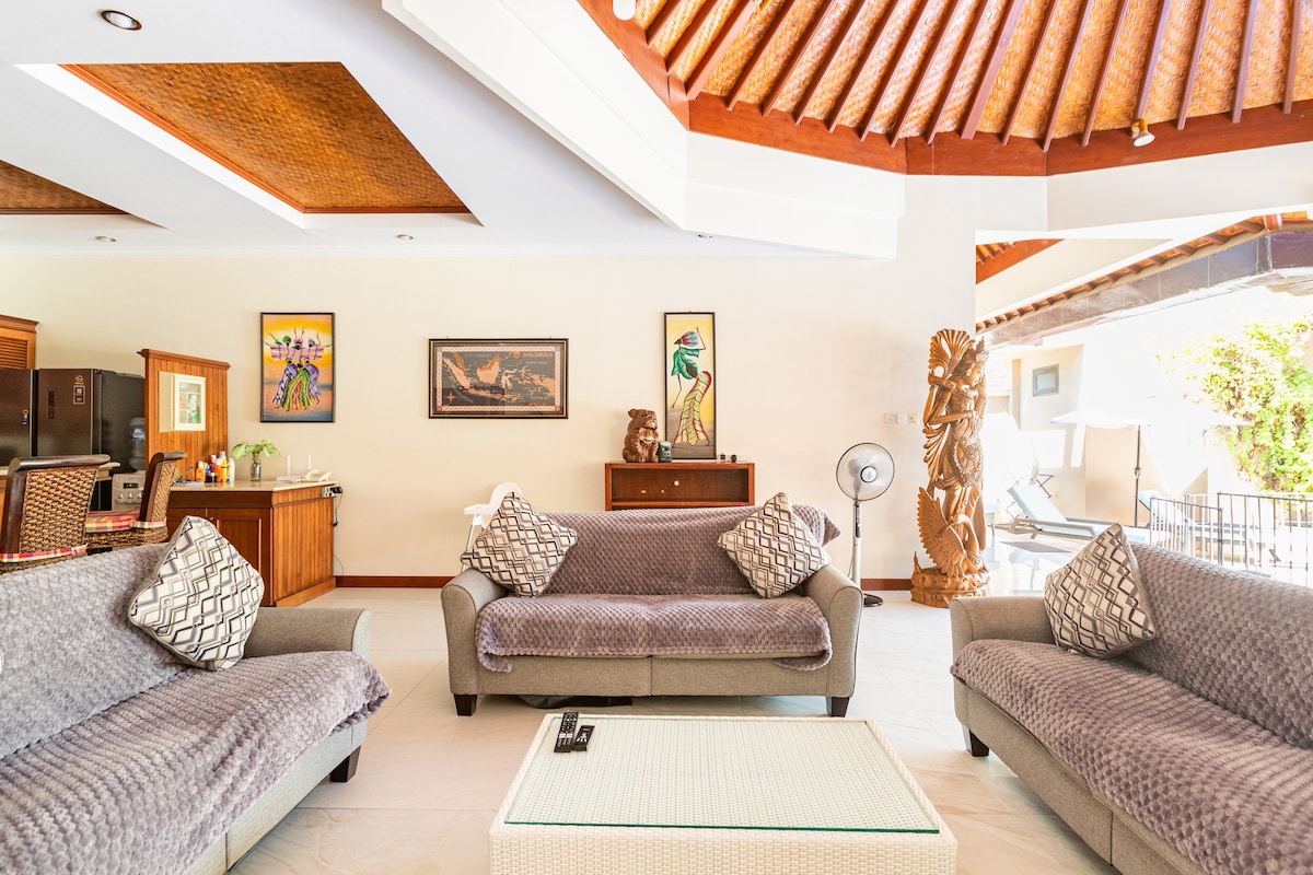An open living area features a high, wooden ceiling with intricate details. Two comfortable sofas are arranged around a light coffee table, accented by decorative cushions. Artwork adorns the walls, with a glimpse of outdoor space visible through large glass doors.