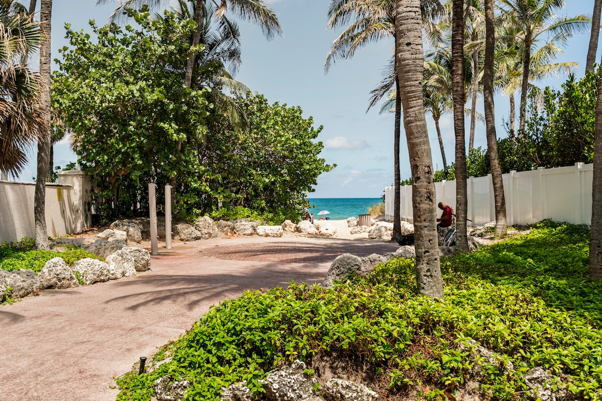 A scenic pathway lined with palm trees leads to a secluded beach. Lush greenery envelopes the area, while soft sandy trails are visible in the soft sunlight. The ocean is visible in the distance, with beach chairs set up along the shoreline.