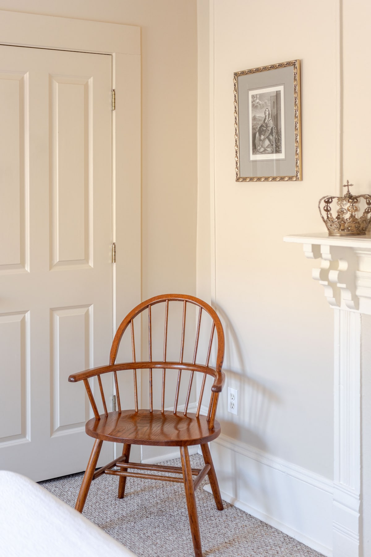 A wooden Windsor chair is positioned in the corner of a softly lit room. It rests next to a closed white door, with a small framed artwork displayed on the light-colored wall above. A decorative crown sits atop the mantelpiece nearby, enhancing the room's charm.