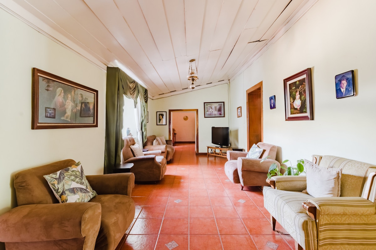 A spacious living area features light-colored walls and a red-tiled floor. Comfortable seating options, including armchairs and a sofa, are arranged to encourage relaxation. Decorative wall art adds visual interest, while natural light filters through large windows, illuminating the space throughout.