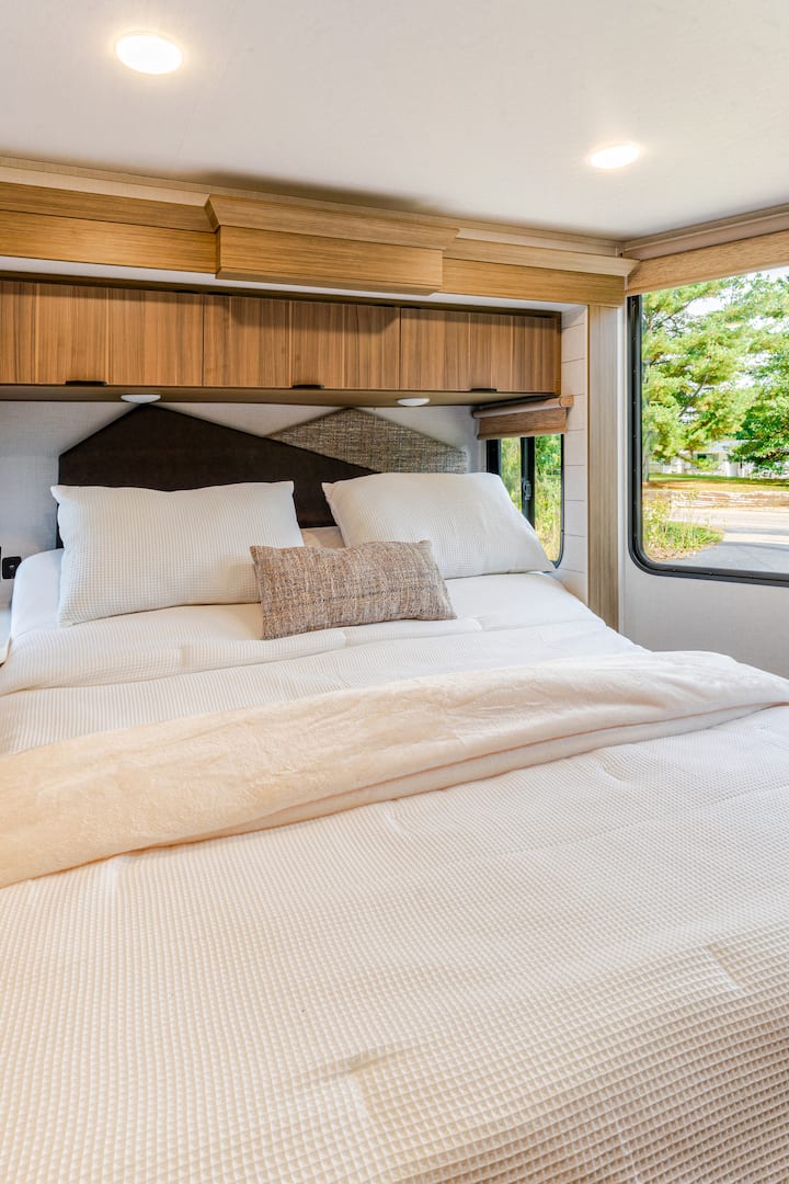 Relax in a cozy retreat featuring a plush queen bed with crisp white linens, soft pillows, and modern wood accents. Natural light filters through large windows, creating a bright yet peaceful space perfect for unwinding after a day by the lake.