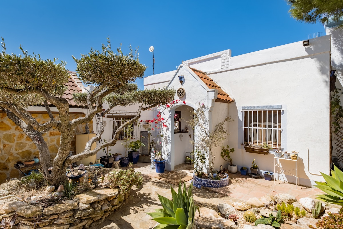 A charming exterior of a white, single-story cottage is surrounded by lush greenery and decorative plants. A stone pathway leads to an arched entrance, framed by flowering shrubs and a manicured tree. The roof is adorned with traditional tiles, complementing the serene atmosphere.