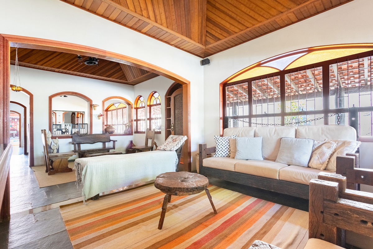 A spacious living area features a wood ceiling and large windows, allowing natural light to fill the room. Comfortable seating is arranged around a low wooden table, with a colorful area rug adding warmth to the space. A dining table is visible in the background.