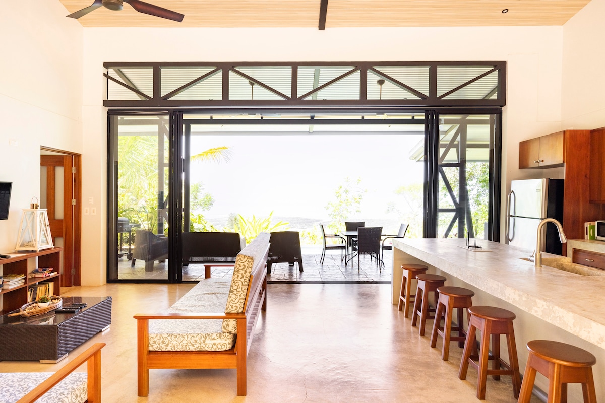 An open living area features modern furnishings, including a spacious sofa adjacent to a concrete kitchen island with wooden bar stools. Large sliding doors provide a view of the terrace and surrounding greenery, allowing natural light to fill the space.