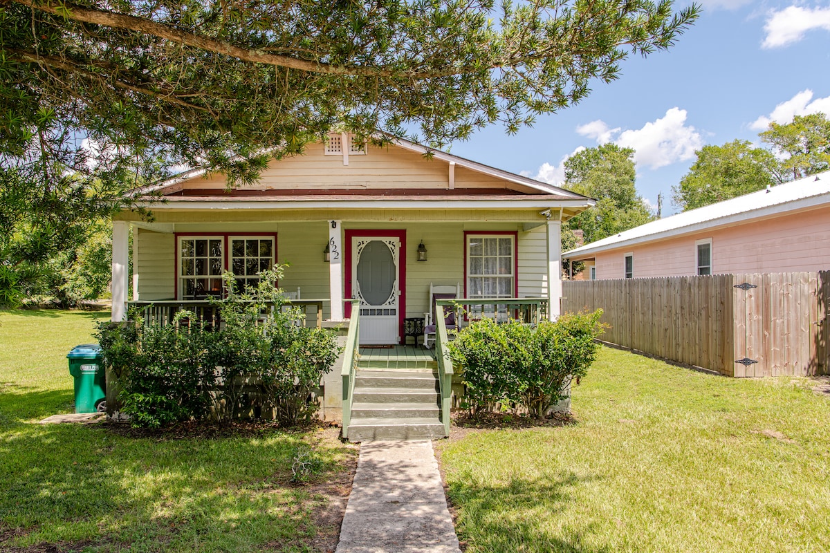 The front of a vintage cottage is framed by a lush yard, showcasing a welcoming porch with a round door and two rocking chairs. A pathway leads to the entrance, flanked by low shrubs and tall grass, providing a cozy and inviting appearance.