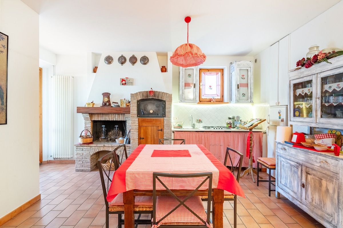 A bright kitchen area features light-colored cabinets and a rustic wooden table set with a red checkered tablecloth. A brick fireplace adds warmth, and decorative plates are displayed on the wall. Natural light enhances the space through a nearby window.