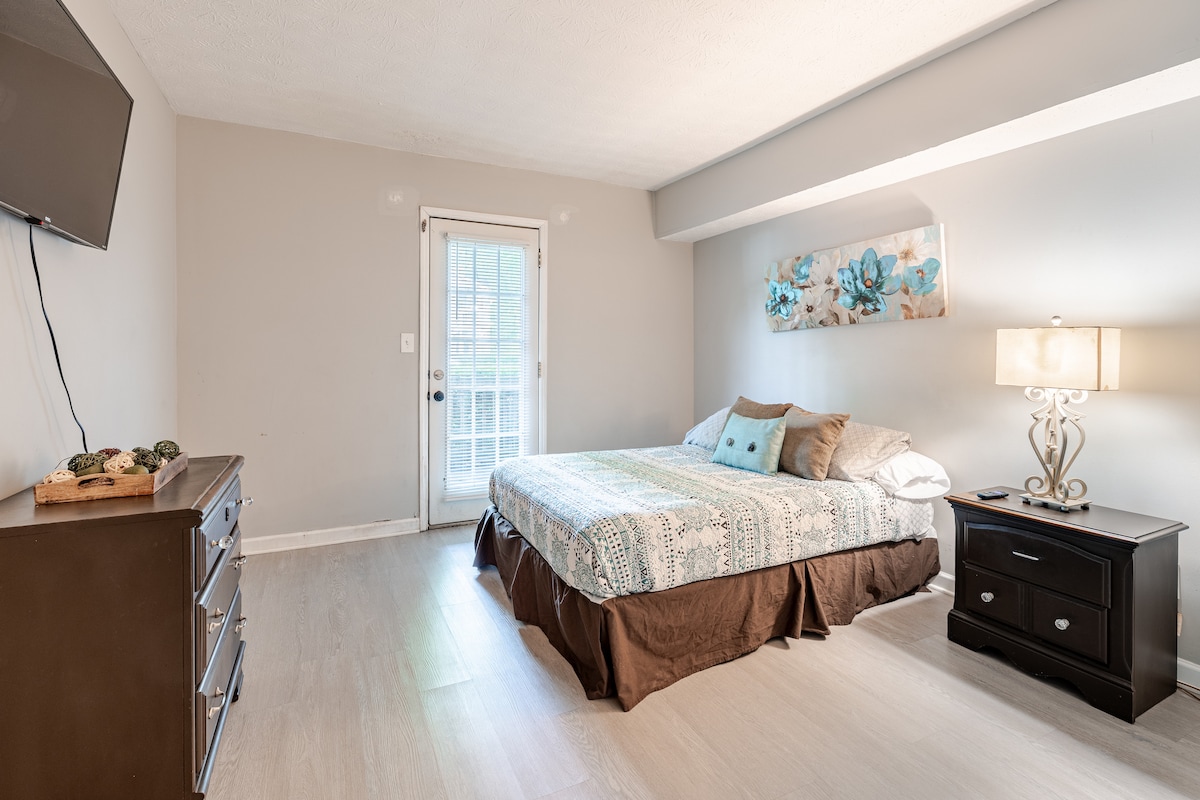 A cozy bedroom is showcased with a spacious bed featuring a patterned duvet. A light-colored dresser and nightstand complement the room. A lamp provides warm lighting beside the bed. A glass door leads to an outdoor area, while artwork adorns the wall above the bed.