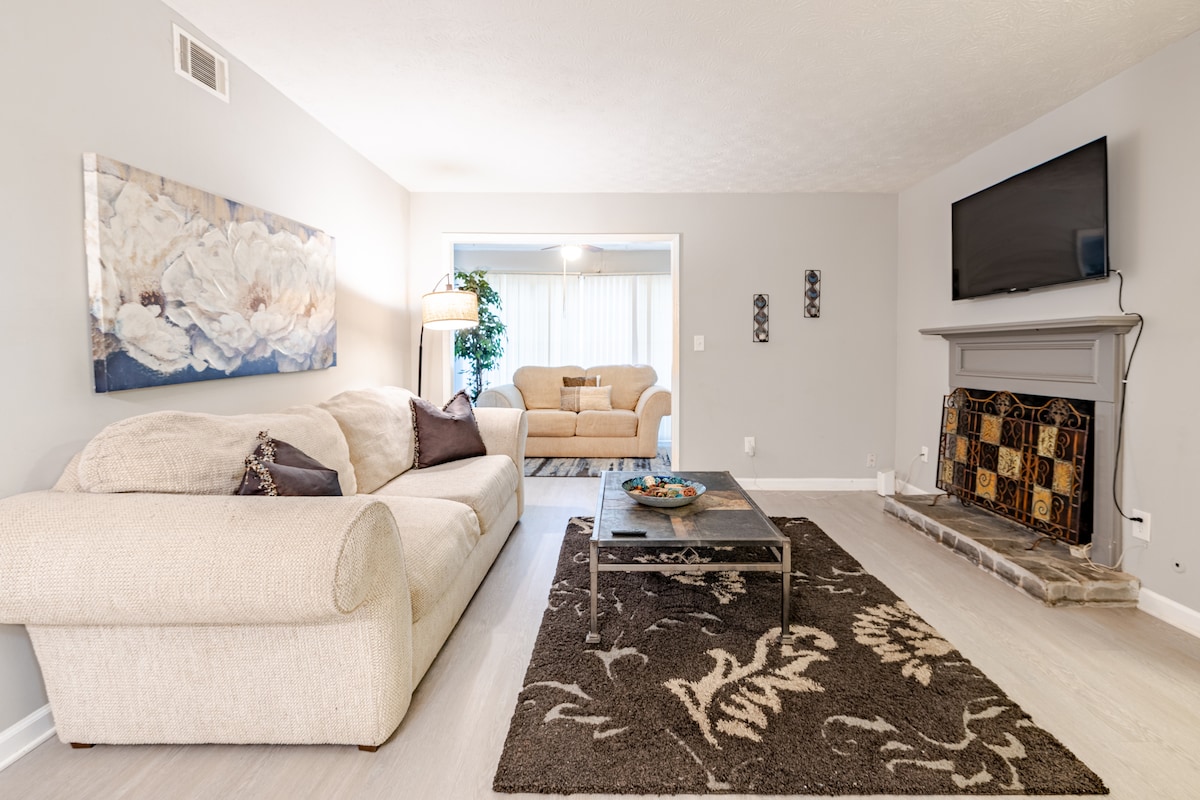 A spacious living area is highlighted by a light-colored sectional sofa and a coffee table, situated on a patterned area rug. A flat-screen TV is mounted on the wall, while a stone fireplace adds a cozy touch. Soft lighting enhances the calm atmosphere.
