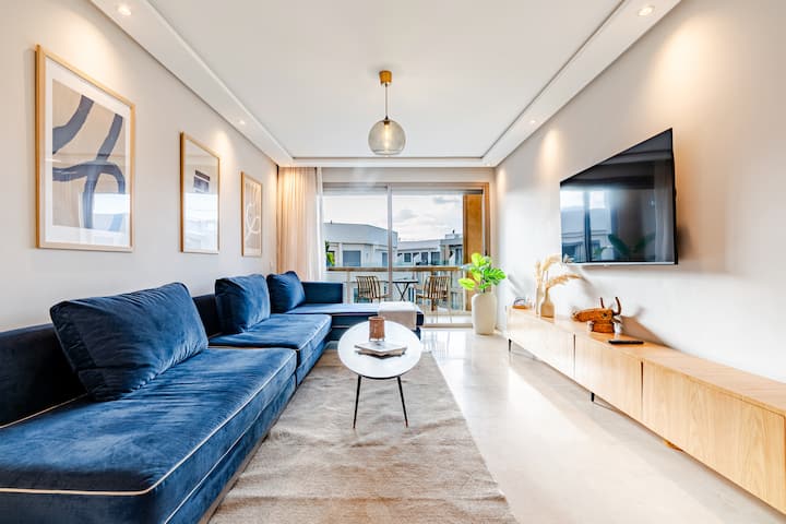 Dnevni boravak svijetlog dizajna s XXL plavim kaučem, zidnim TV-om i drvenim ormarićem, umjetničkim okvirima i velikim kliznim prozorom koji se otvara na namješteni balkon. Svijetli mramorni pod, prirodno svjetlo, idealno za obitelji, prijatelje ili poslovne boravke.