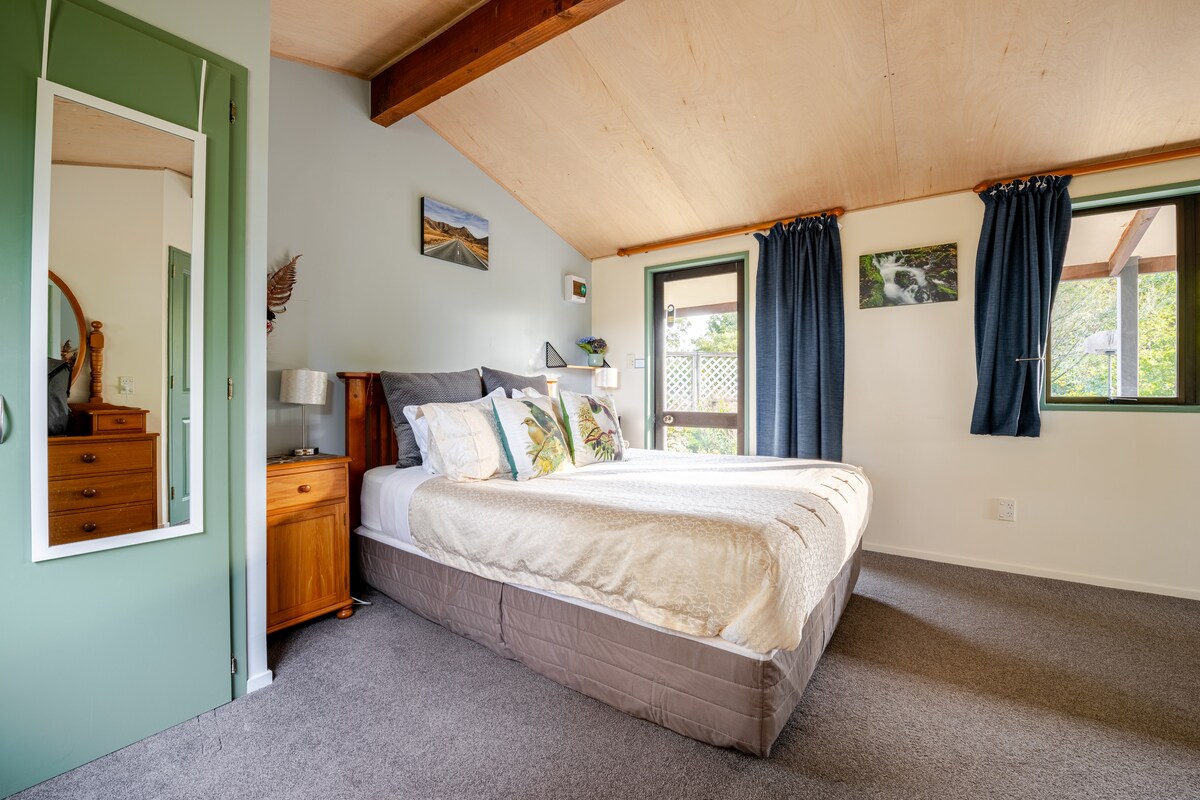 A bright bedroom features a spacious bed with a neutral-colored comforter and decorative pillows. Large windows allow natural light to enter, complemented by dark curtains. A wooden nightstand and dresser add warmth, while a full-length mirror reflects the cozy atmosphere of the room.
