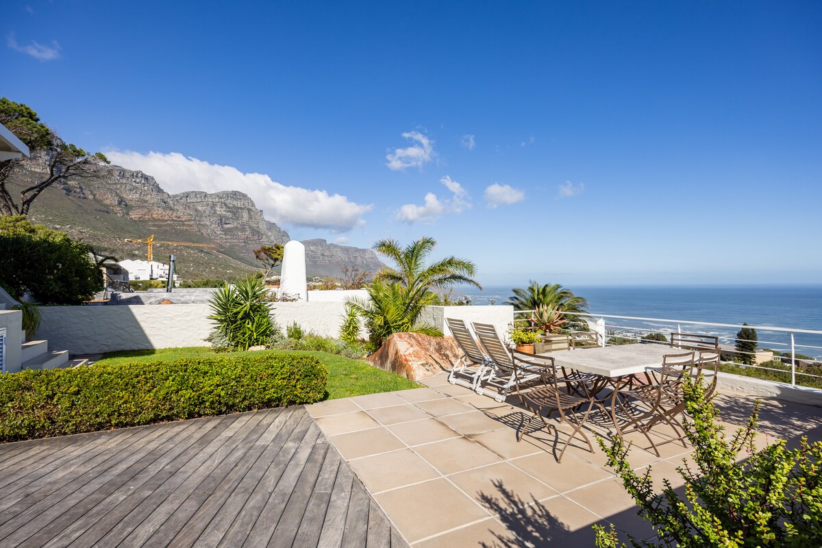 A sunlit outdoor terrace is shown, featuring a large rectangular table surrounded by several chairs. Lush greenery and ornamental plants frame the space, while majestic mountains rise in the background, leading down to the expansive ocean below, under a clear blue sky.
