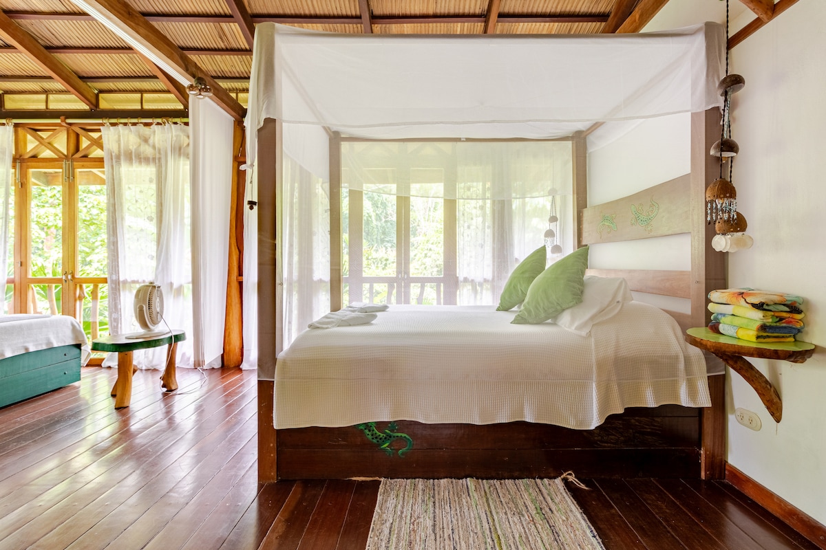 A spacious sleeping area features a wooden bed with a canopy draped in sheer fabric. Two green pillows accentuate the white bedding. Large windows allow natural light to fill the room, while wooden floors complement the rustic design.