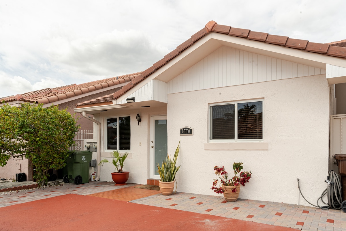 The exterior view of the home is presented, featuring a light-colored facade and a welcoming entrance. Potted plants are positioned near the door, and a red pavement leads up to the house. An assortment of greenery is visible, adding to the inviting atmosphere.