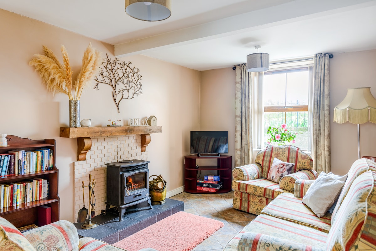 A cozy living room features a traditional log burner on a slate hearth, flanked by light-colored walls and natural decor. A bookshelf holds an array of books, while comfortable sofas with striped upholstery invite relaxation. Natural light filters through the window adorned with floral curtains.