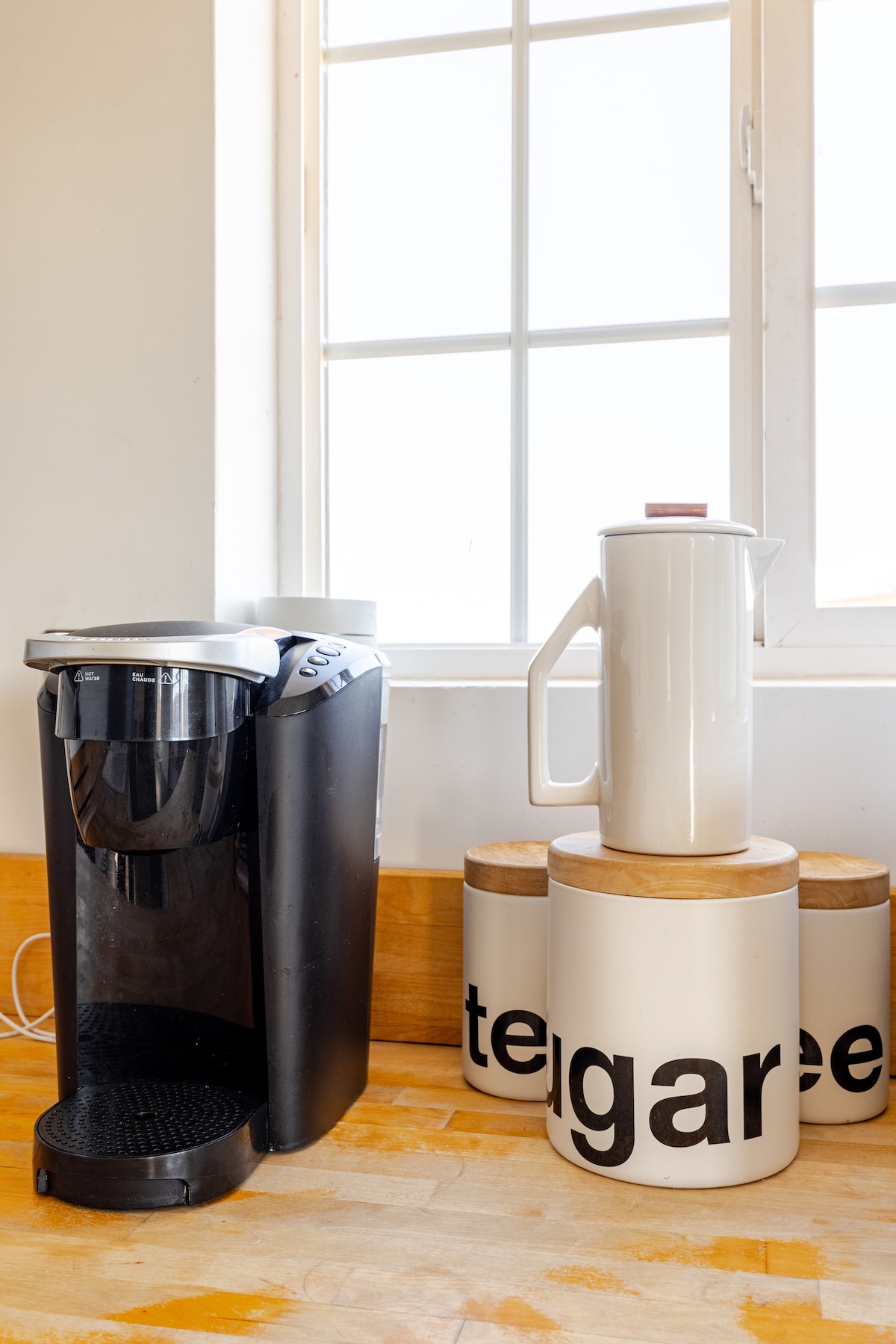 A modern coffee maker is positioned beside a sleek French press, resting atop circular wooden coasters. Light enters through a large window, illuminating the workspace. Decorative canisters labeled 'tea' are arranged on the countertop, enhancing the kitchen's functionality.