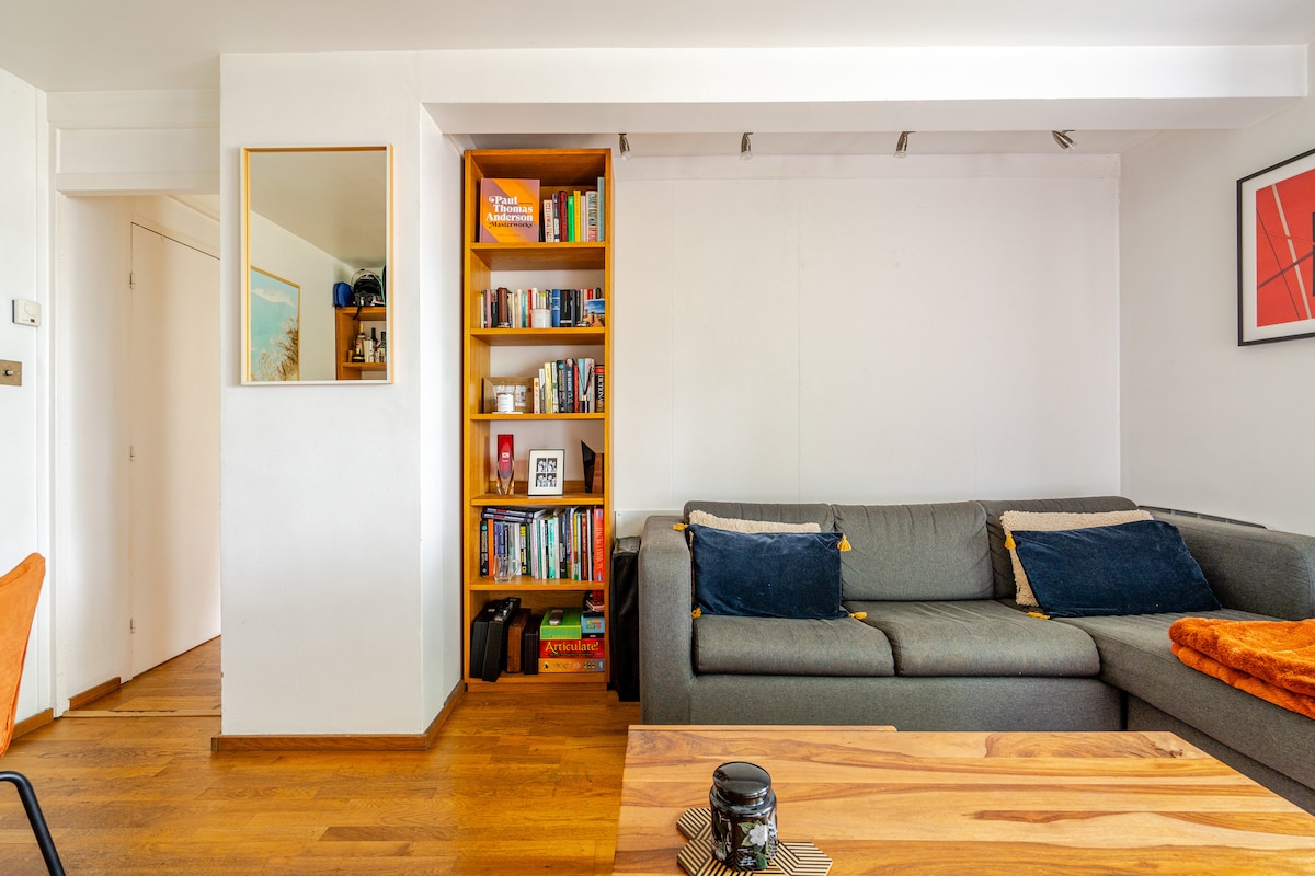 A spacious living area features a comfortable gray sofa adorned with cushions. A wooden coffee table sits in front, while a bookshelf filled with various books is visible to one side. A mirror hangs on the adjacent wall, reflecting the light-colored decor.