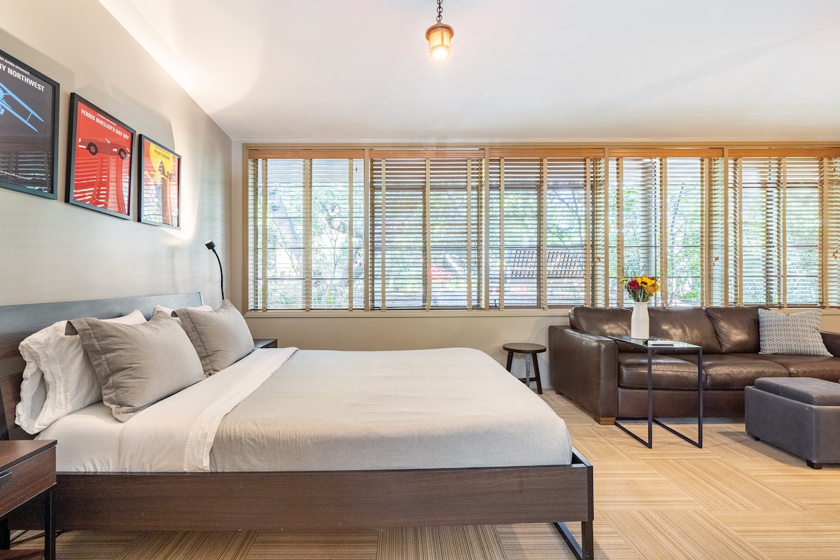 A spacious sleeping area features a large bed with neutral-toned bedding. Natural light is filtered through restored casement windows, creating a bright ambiance. A comfortable leather couch and a side table are positioned nearby, complemented by decorative artwork on the walls.