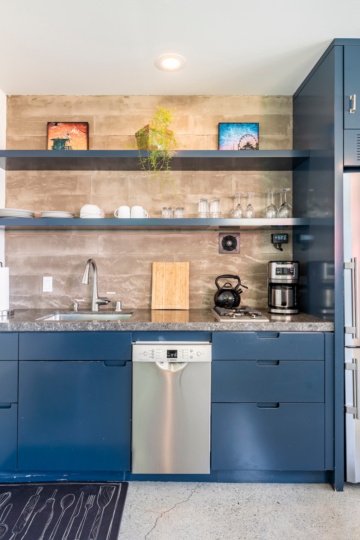 A modern kitchen features navy blue cabinets and stone countertops, offering an efficient layout. Glassware is neatly arranged on open shelves above the sink, while a coffee maker and kettle are positioned on the counter. Two pieces of artwork add a touch of color to the wall.