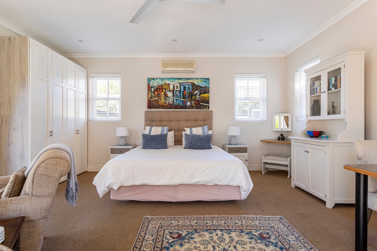 A spacious bedroom is showcased with a large bed adorned with decorative cushions. Soft light filters through two windows, illuminating the neutral walls and a piece of art above the headboard. Storage cabinets and bedside tables provide convenience, while a cozy armchair invites relaxation.