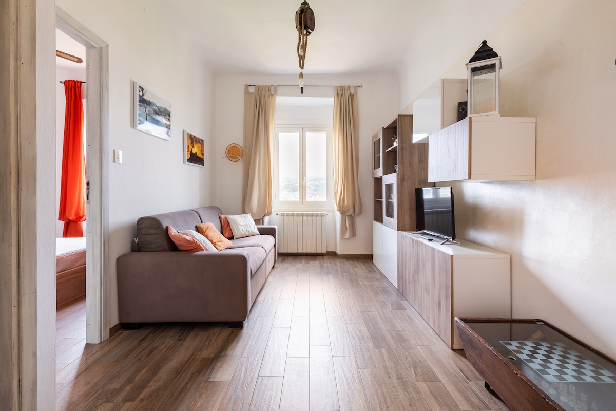 A light-filled living area features a comfortable sofa with decorative pillows. Dark wood flooring extends throughout the space, complemented by a modern TV unit and shelving. Large windows offer views of the surrounding landscape, and soft drapes frame the natural light entering the room.