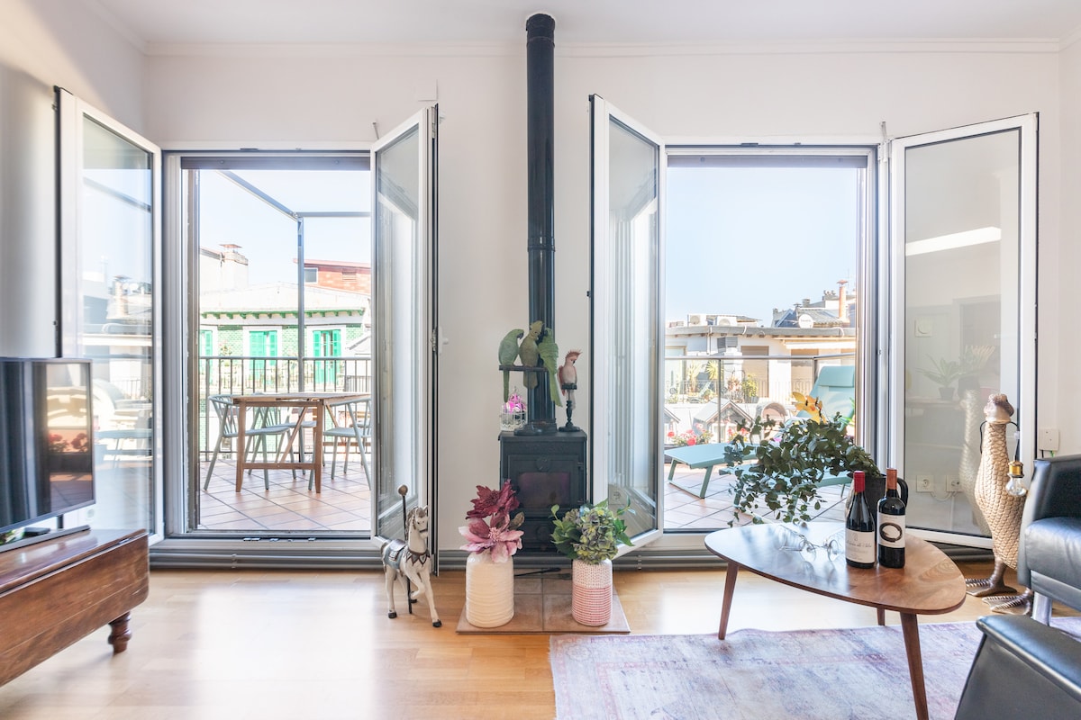 A bright interior space showcases large open windows that extend to a terrace view. A cozy sitting area includes a wooden table with a wine bottle and two glasses. A small fireplace adds warmth, complemented by decorative plants beside it.