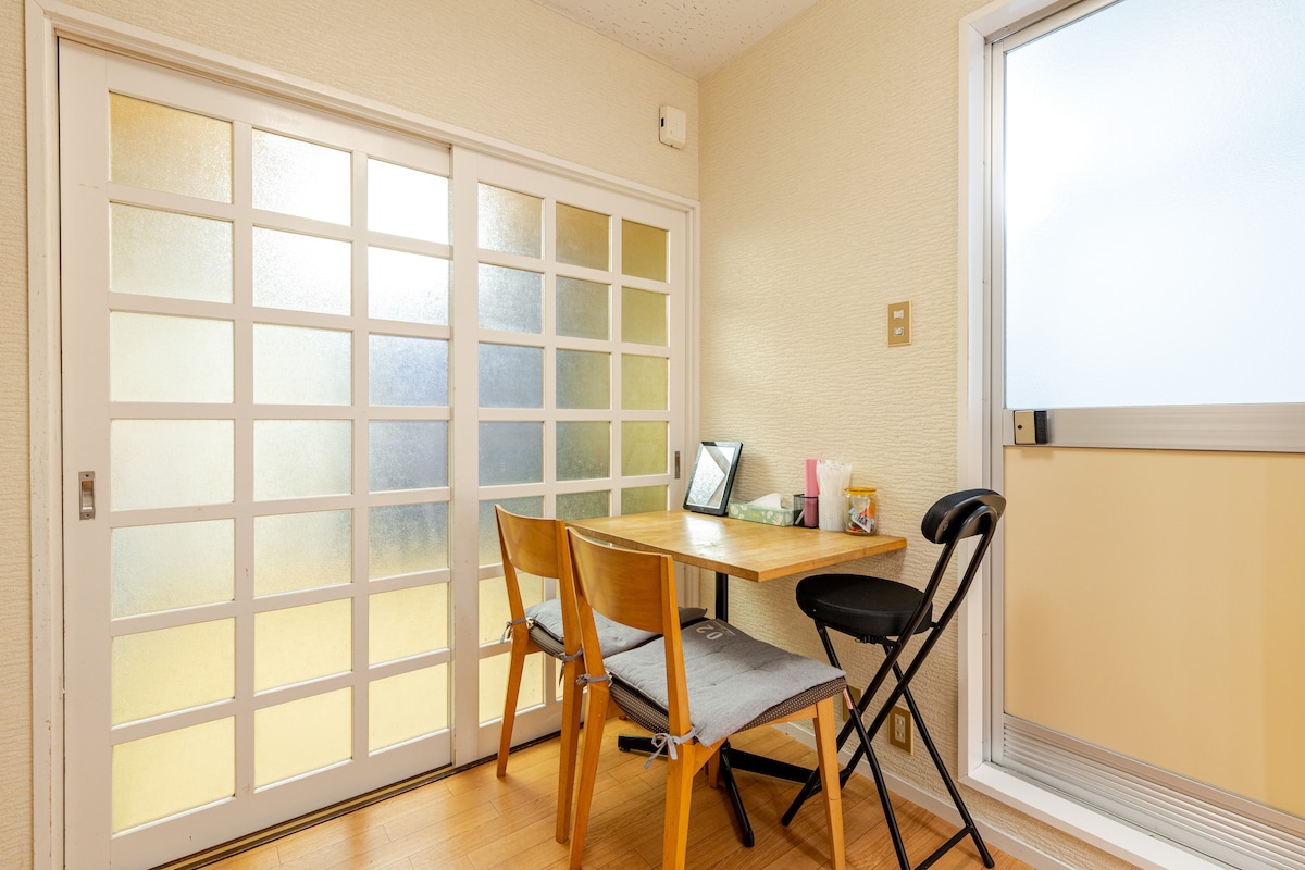 A small dining area features a wooden table accompanied by two chairs and a folding stool. Large glass doors permit natural light, and a window offers an outdoor view. A reflection and soft patterns on the doors enhance the space's simplicity.