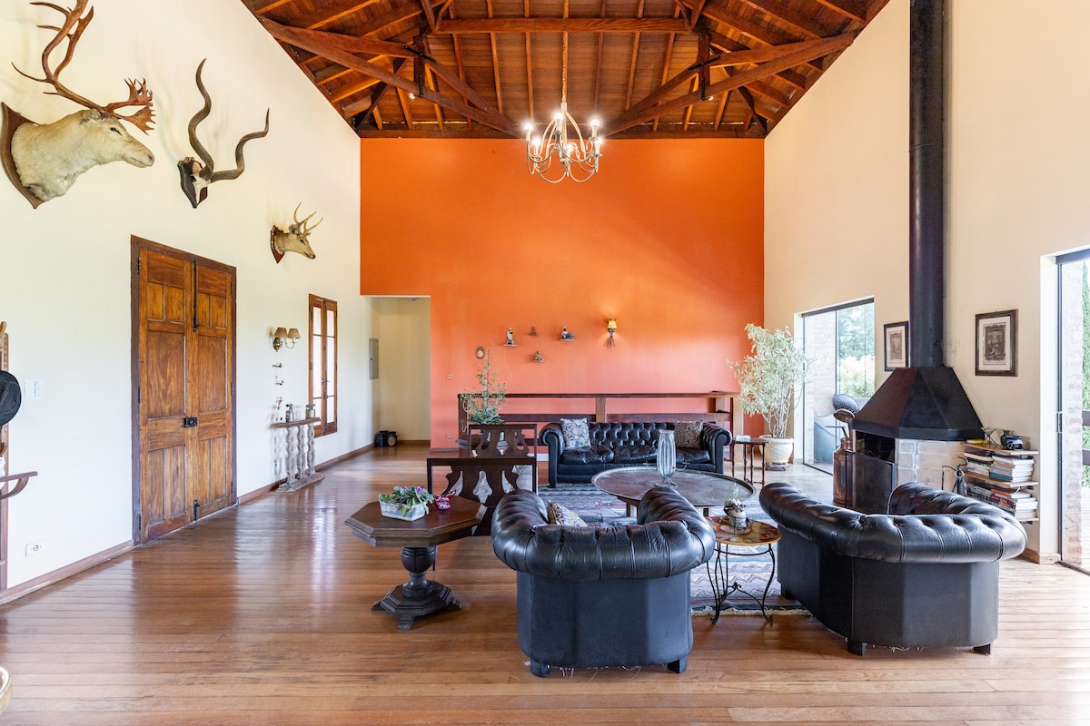 A spacious living area features a high wooden ceiling and large windows that provide ample natural light. Elegant leather sofas are arranged around a central table. A vibrant orange wall complements the cozy atmosphere, while decorative elements like mounted art are subtly displayed.