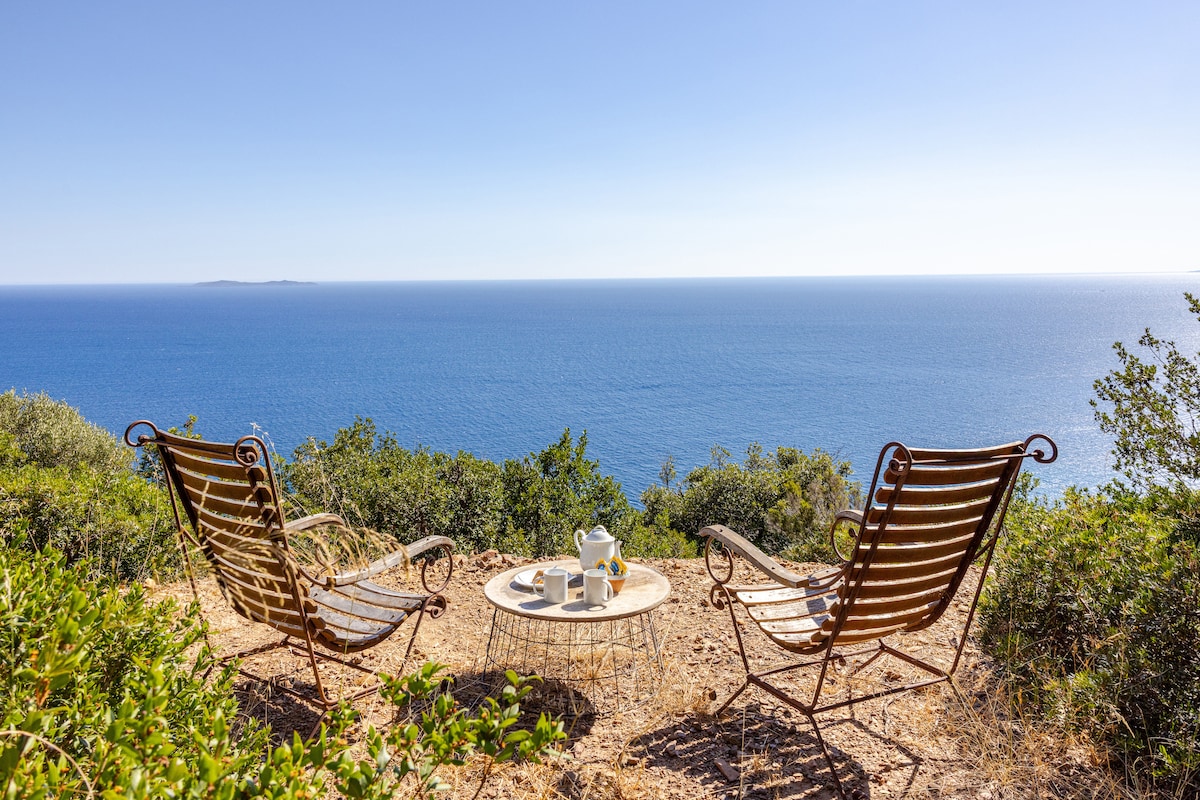 Two rustic metal chairs are positioned on a hillside overlooking the vast Mediterranean Sea. A small table is set between them, displaying a teapot and cups. Lush greenery surrounds the seating area, enhancing the serene coastal atmosphere.