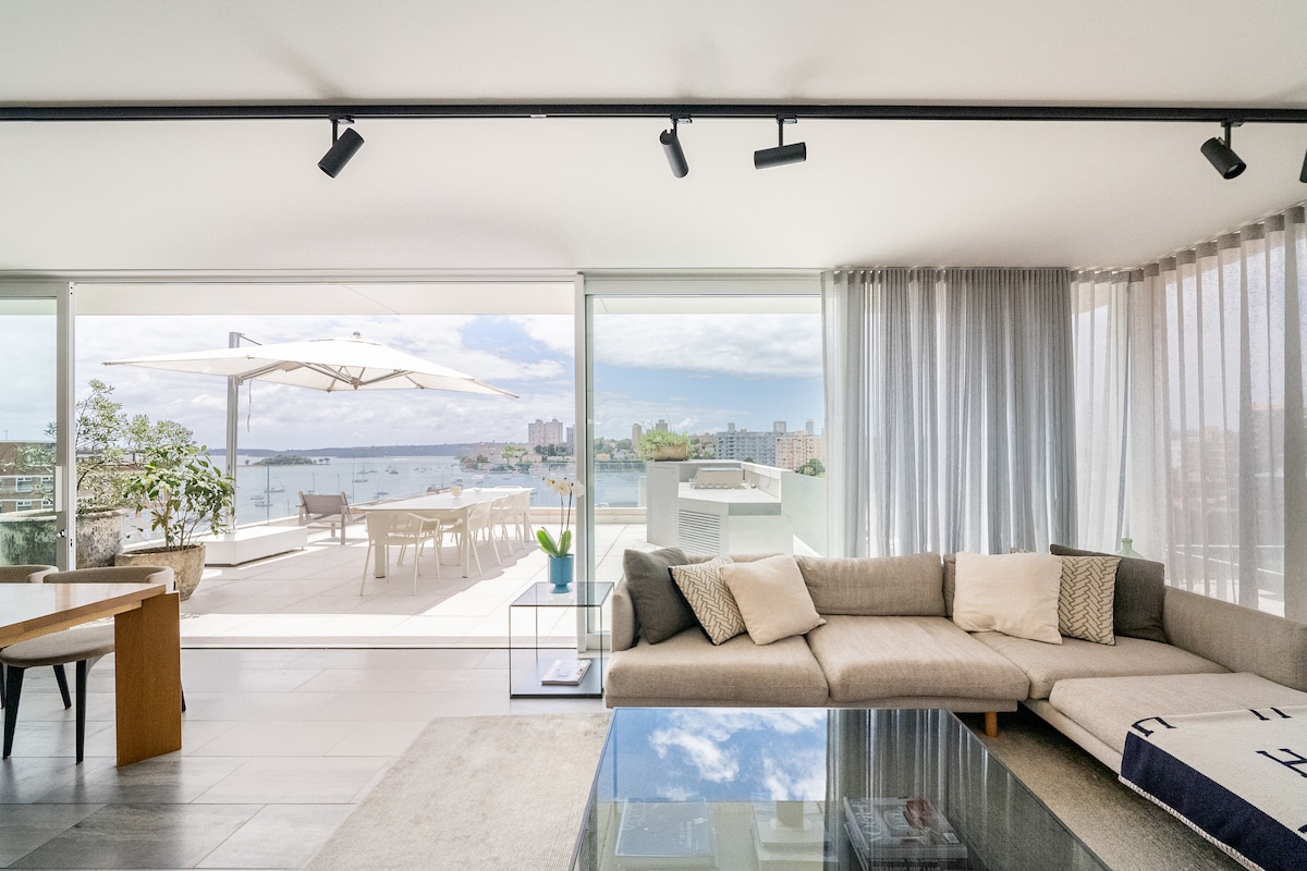 A spacious living area features a large sectional sofa adorned with neutral pillows. Natural light floods the space through expansive glass doors that open to a terrace with panoramic views. A sleek coffee table sits on the tiled floor, complementing the contemporary decor.